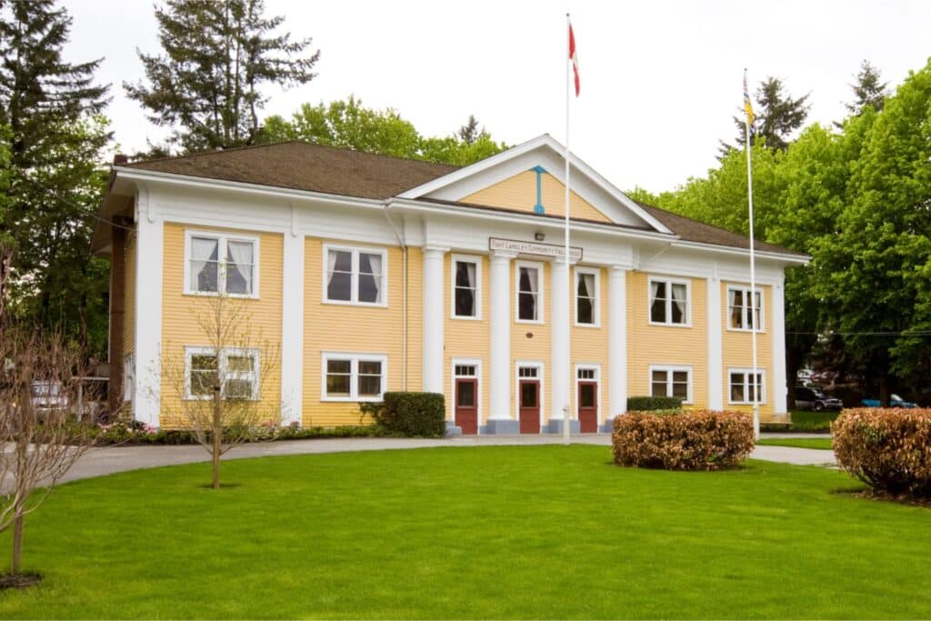 Fort Langley Community Hall is a two storey yellow building with colonial style white columns on its front, seven windows across the top storey, and four storeys and three doors across the bottom. The doors are red and trimmed in white; the windows are trimmed in white. In front of the hall is a semi-circle of grass with a semi-circle pathway. There is a B.C. flag on the flagpole on the left, and there is a Canada flag on the flagpole on the right.