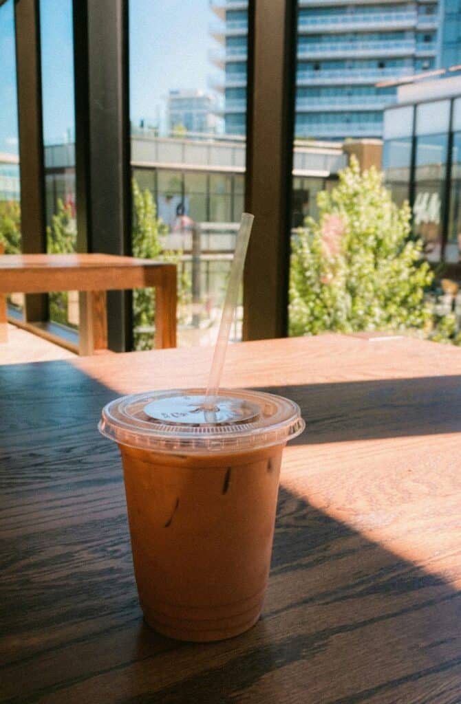 Iced coffee in a clear plastic cup sitting on a table next to a window that looks at buildings and towers and trees on a sunny day in the neighbourhood of Brentwood in Burnaby, B.C., Canada.