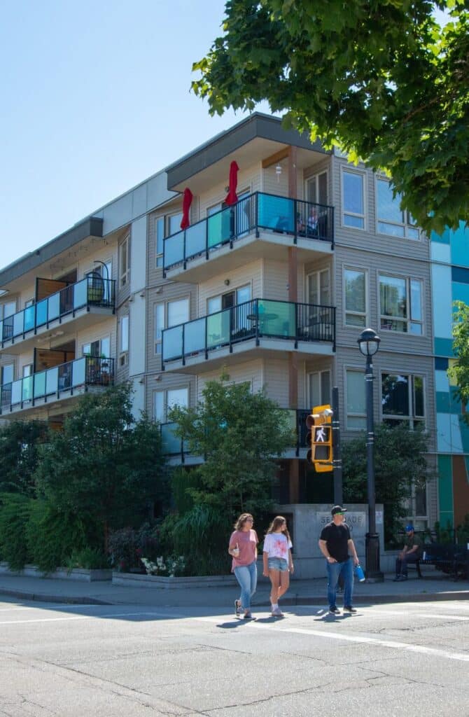 Modern four-story apartment building with balconies, red umbrellas, and pedestrians crossing in front on a sunny day in downtown Langley, B.C., Canada.