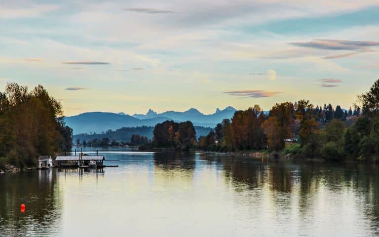A north facing image of the Fraser River looking at a mountain range with trees on both sides of the river with blue sky, scattered white clouds and a full moon in Langley, B.C., Canada