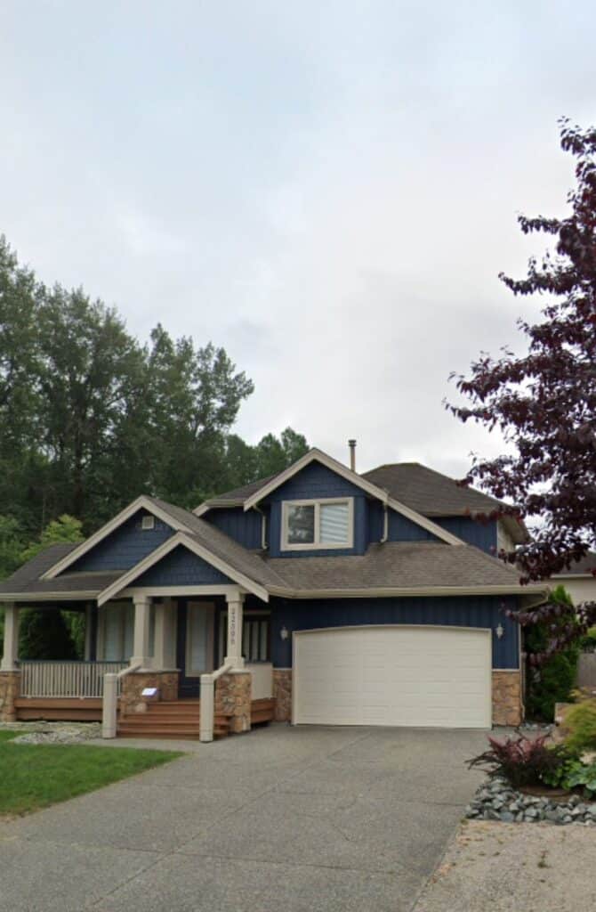 Two-story blue house with a front porch, white garage door, and surrounding trees and landscaping in Langley, B.C. Canada.