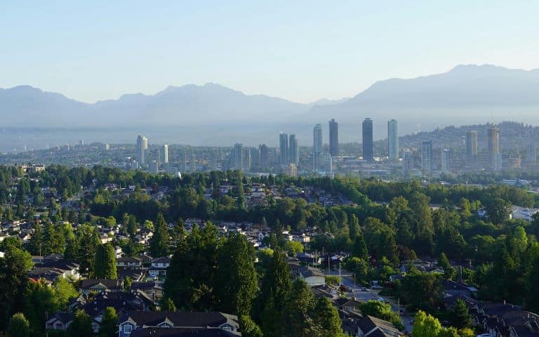 Scenic photo of green trees, residential area and city towers in Burnaby, B.C., Canada against a backdrop of mountain range and blue sky.