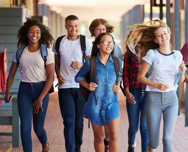A group of six high school teenagers walking and smiling with their backpacks on at school wearing jeans and short sleeved t-shirts and one girl wearing a jean dress.