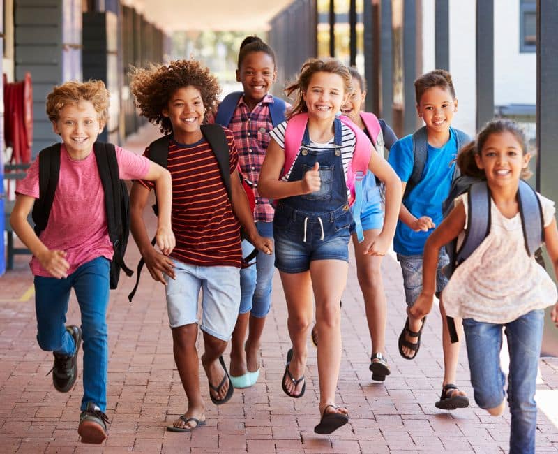 A group of smiling school aged children running with their backpacks on at school wearing summer clothes.