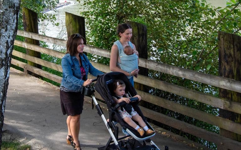 A woman walking pushing a stroller with a girl toddler next to a woman walking with a baby carrier on her front on a trail path with trees and a wooden fence between them and the Fraser River in Fort Langley, B.C. Canada.