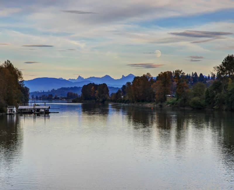 A north facing image of the Fraser River looking at a mountain range with trees on both sides of the river with blue sky, scattered white clouds and a full moon in Langley, B.C., Canada