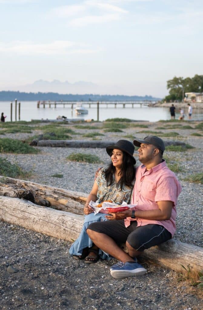 A man and woman sitting on a log on the beach with baskets of fish and chips on their laps looking out at the water against a background of a boat, pier and other people along the beach in Surrey, B.C., Canada.