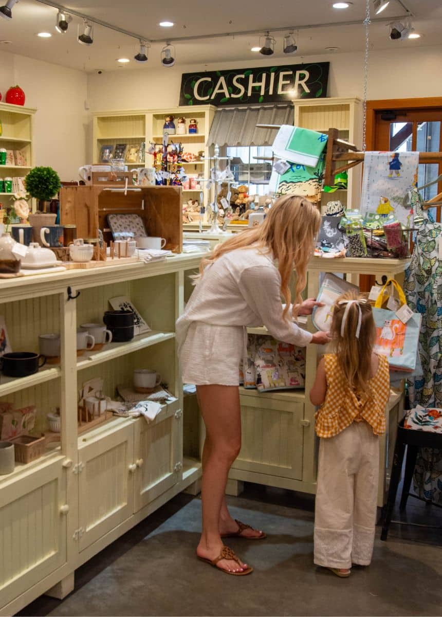 A woman with long blonde hair wearing a long-sleeve white shirt, white shorts and brown sandals standing next to a young girl with long blond hair with a white bow, yellow tank top and white pants. Both facing backwards to the camera in a farmhouse style store in Langley, B.C., Canada.
