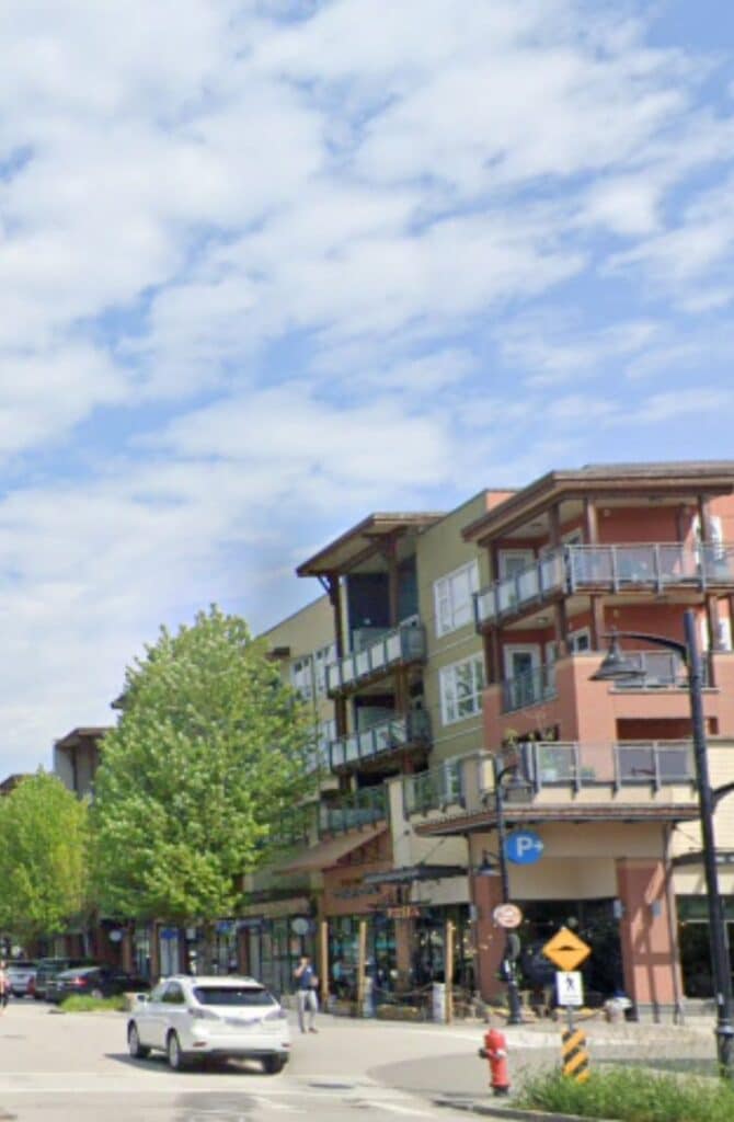 Urban street view with multi-story buildings, trees, parked cars, and a white car driving under a sunny and partly cloudy sky in Langley, B.C., Canada.