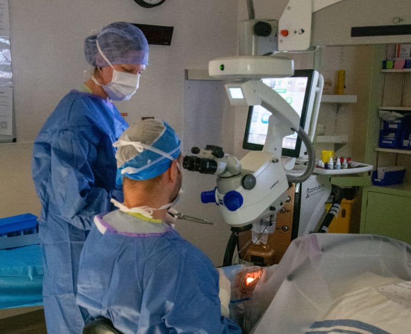 A male doctor and female nurse in blue scrubs, gloves, mask and cap in an operating room operating on an eyeball. Doctor is sitting looking through medical equipment and the nurse in standing handling tools while the patient lies flat on a gurney at Langley Memorial Hospital in Langley, B.C., Canada.