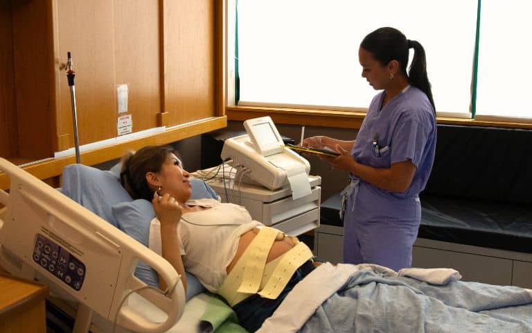 A female medical professional in blue hospital scrubs holding a clipboard and pen standing next to a pregnant woman who is lying on a bed in a hospital room hooked up to a fetal monitor at Langley Memorial Hospital in Langley, B.C., Canada.