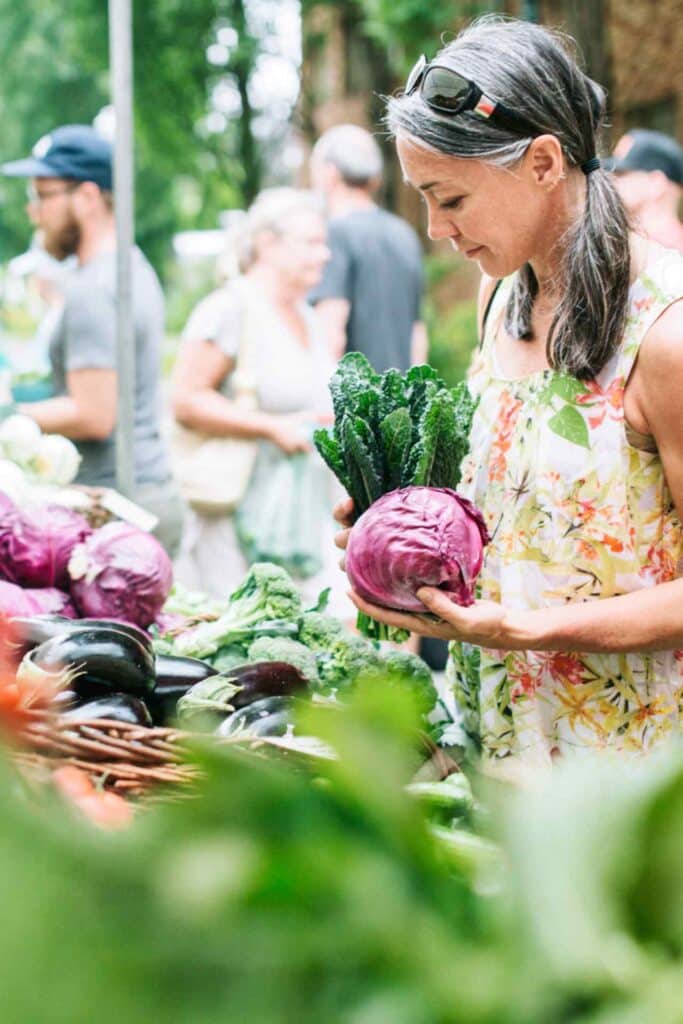 A woman holding kale and cabbage at a farmers' market stall with fresh vegetables and shoppers in the background.
