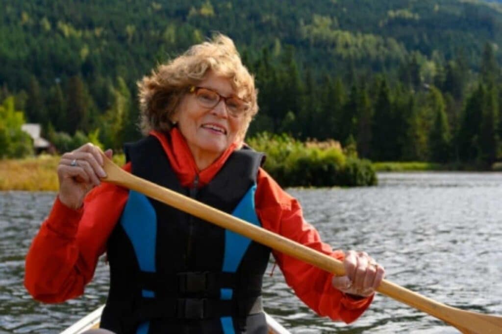 Smiling older woman in an orange jacket and blude and black life jacket paddling in a canoe with hill of trees behind.
