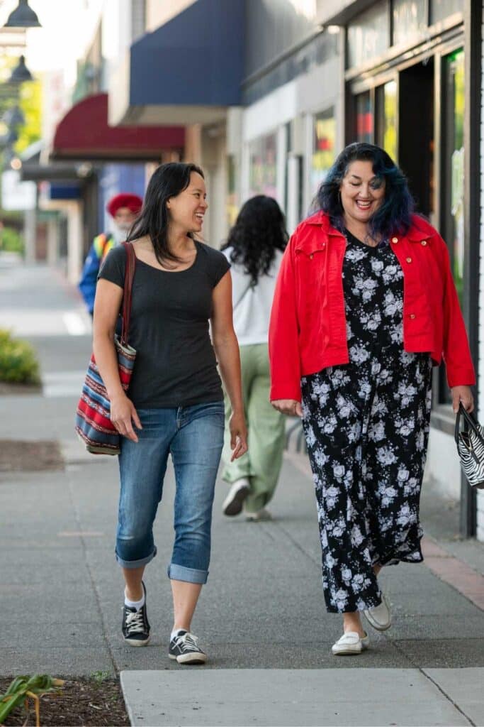Two women walking on a sidewalk in a shopping area.