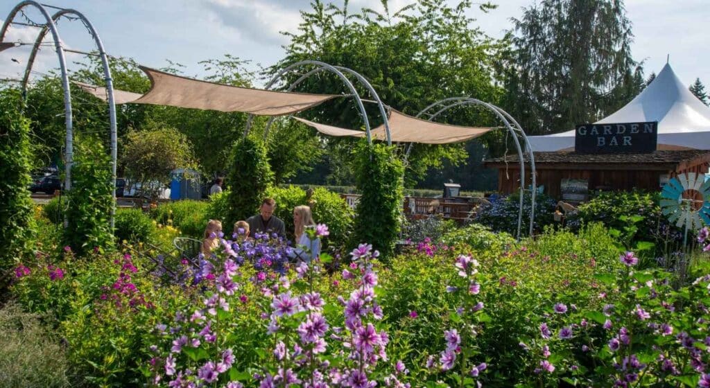 People seated under shaded arches in a garden at Krauss Berry Farm with purple flowers, near a wooden 'Garden Bar' structure and surrounded by greenery in Langley, B.C., Canada